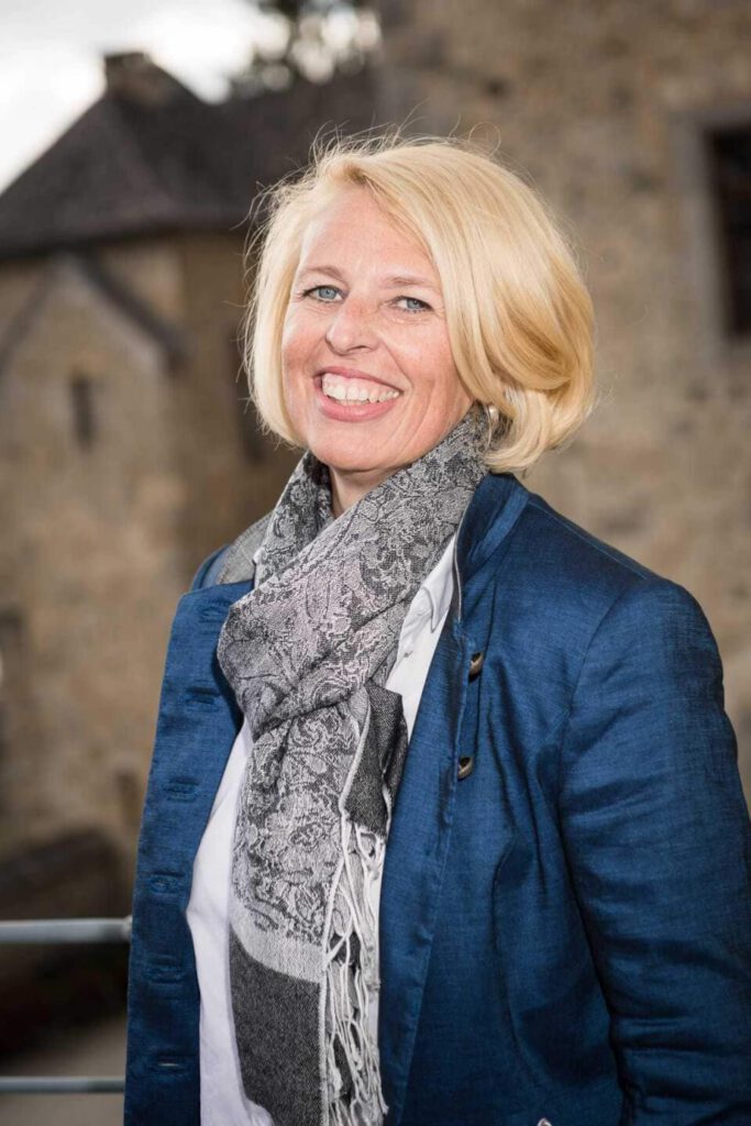 Doris Wallner lächelt in einem blauen Jacket mit Schal, vor einer historischen Kulisse.